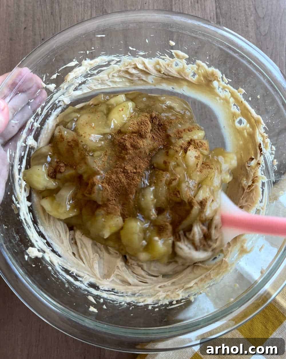 A kitchen scene demonstrating the process of stirring apple pie filling into the prepared cream cheese mixture using a spatula within a mixing bowl. The apples are still largely intact, ensuring a chunky texture in the final dip.