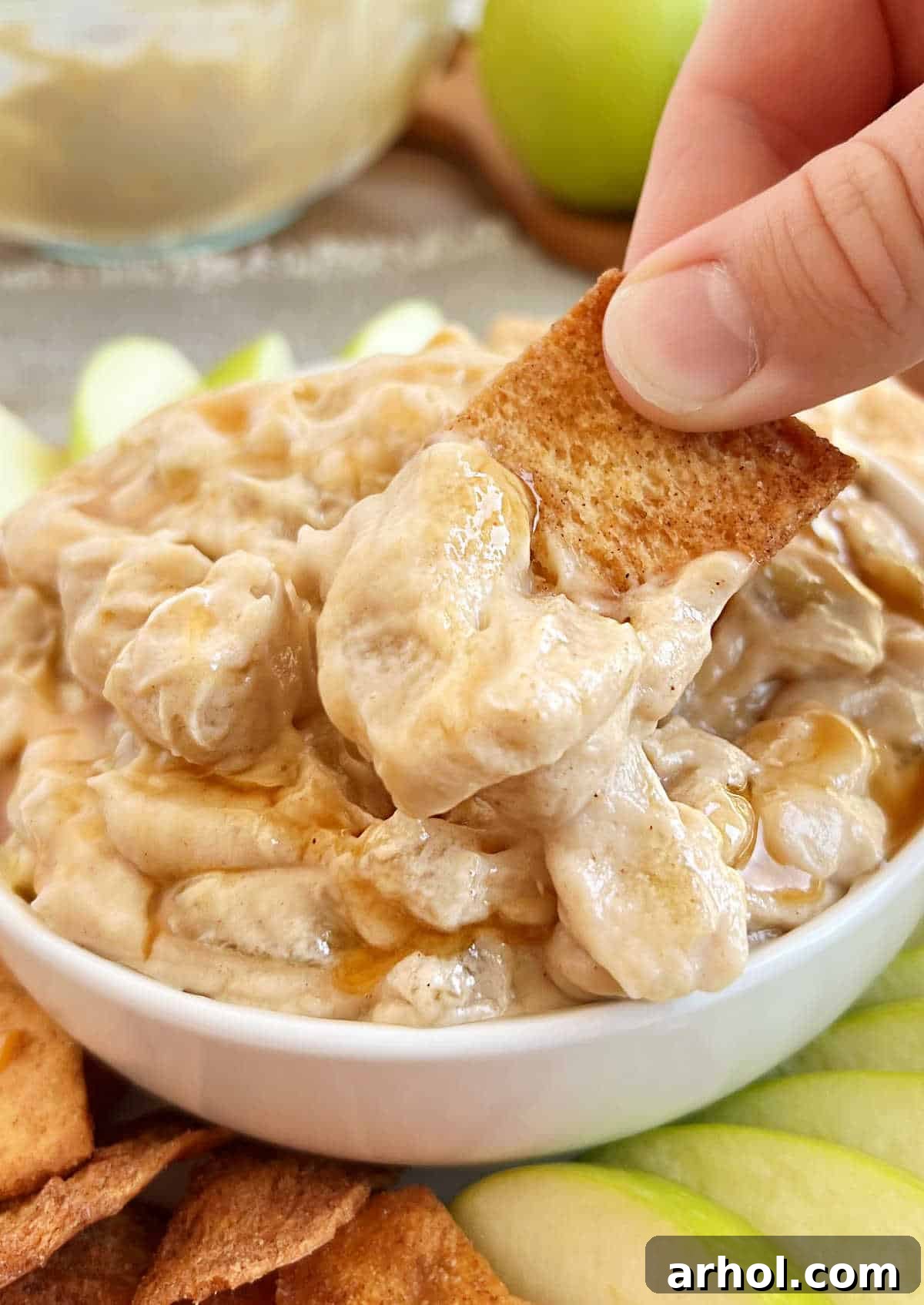 A single, perfect scoop of creamy apple pie dip generously placed on a crisp cinnamon sugar pita chip, artfully positioned against a backdrop of fresh apple slices and more chips, signifying it's ready for a delicious bite.