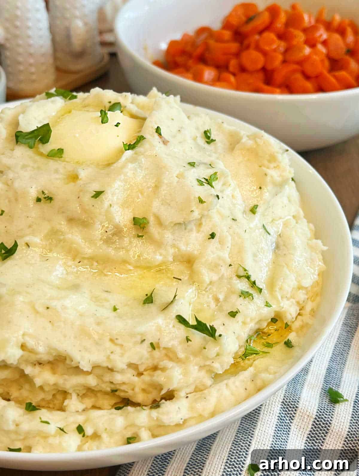 Cream cheese mashed potatoes with melted butter and fresh herbs in a white serving bowl, ready to be enjoyed.