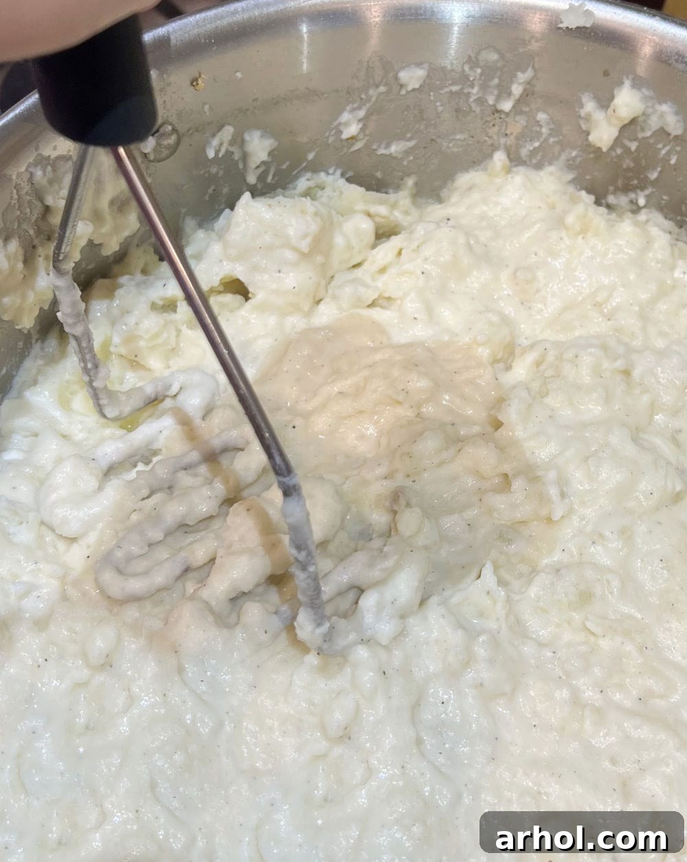A potato masher actively mashing boiled potatoes with cream cheese and butter in a pot, creating a creamy texture.