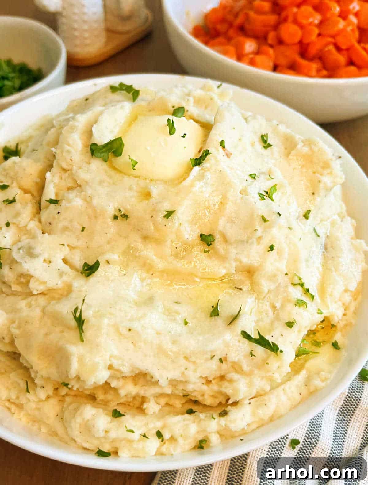 A large serving bowl filled with creamy mashed potatoes, adorned with melted butter and fresh parsley, sitting on a rustic wooden table.