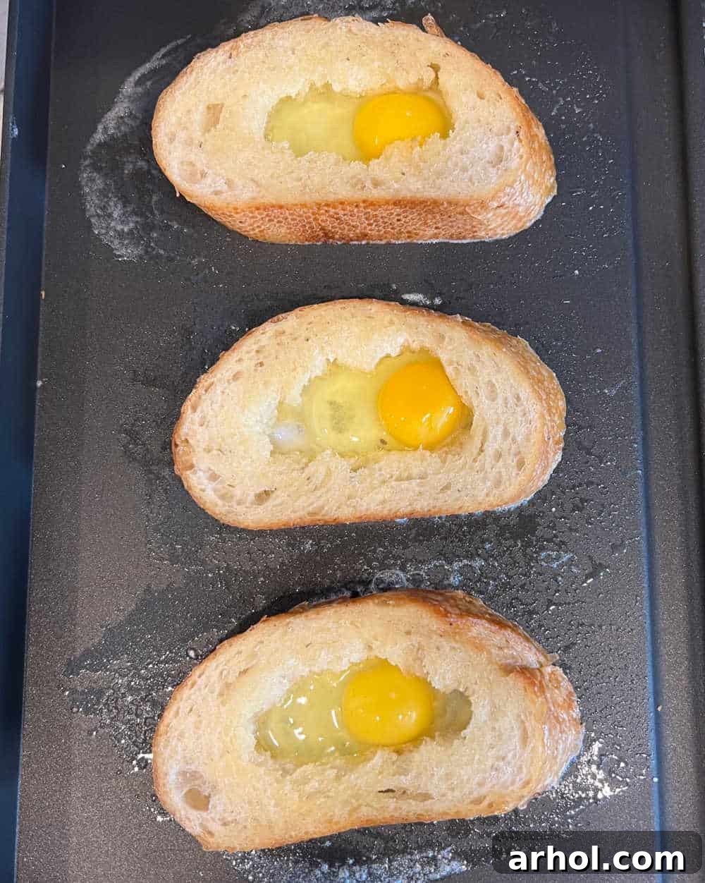 Pieces of bread with eggs cracked into the middle, cooking on a griddle until golden brown.