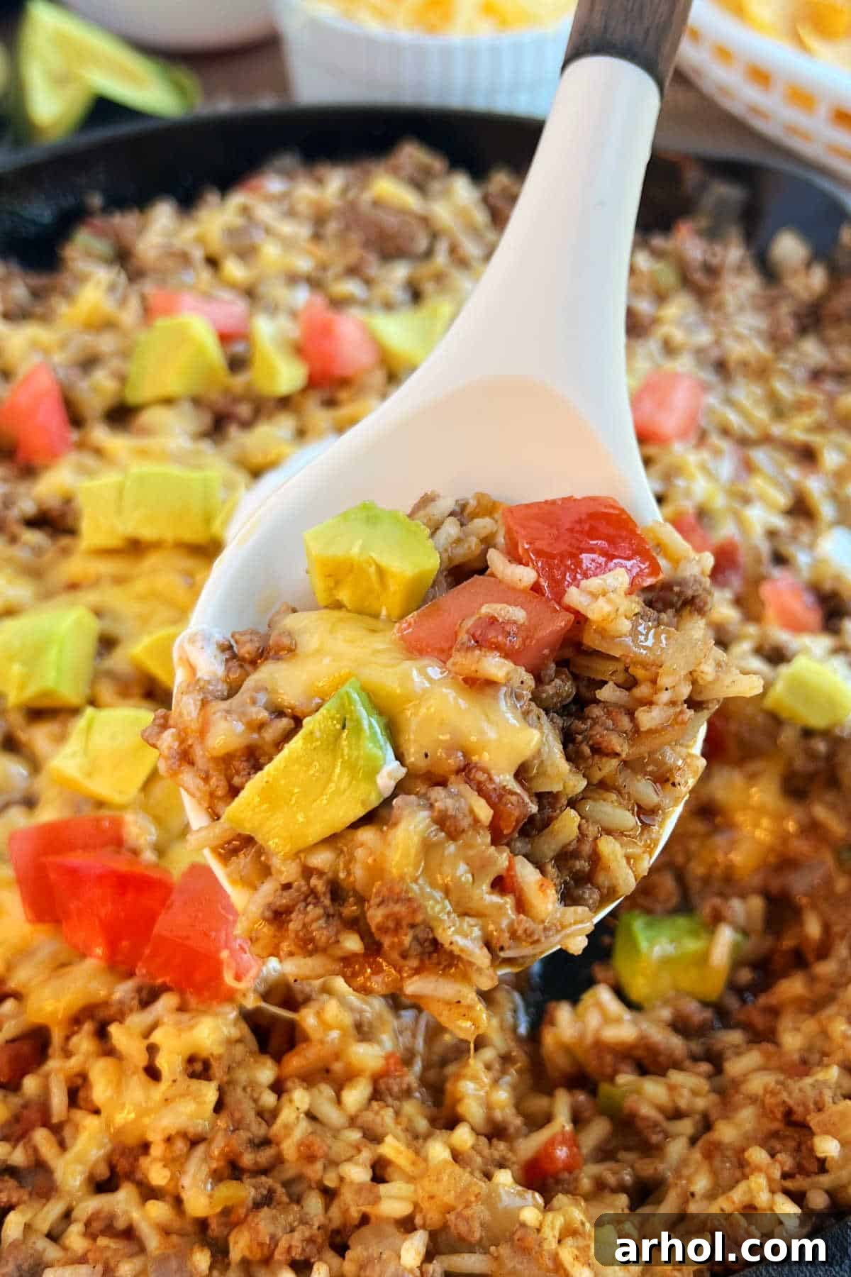 Close-up of a serving spoon scooping cheesy skillet taco rice, garnished with fresh tomatoes and creamy avocado.