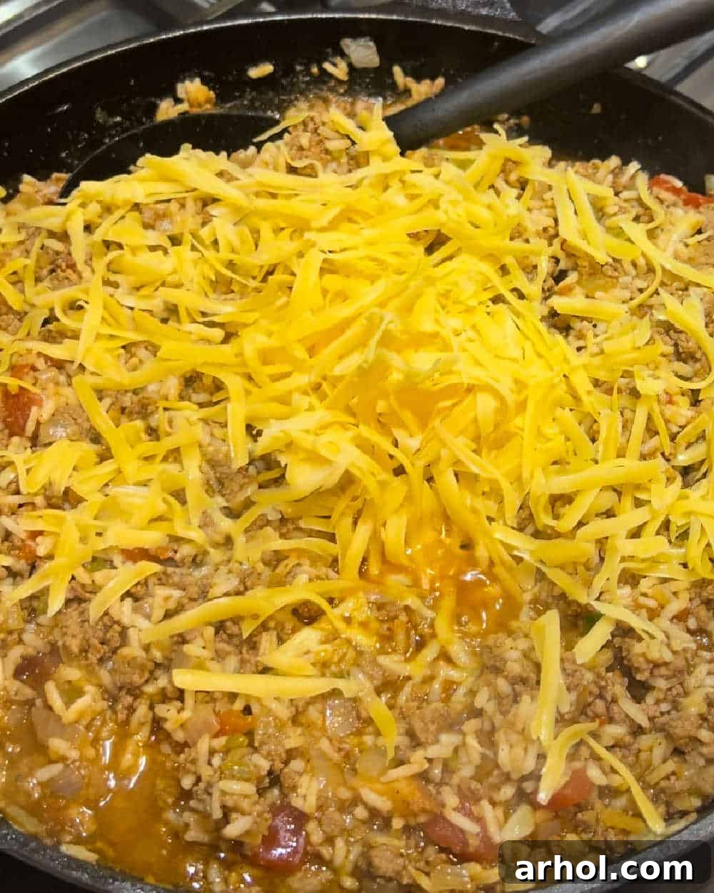 Melted shredded cheddar cheese being stirred into the cooked ground beef and rice mixture in a skillet.