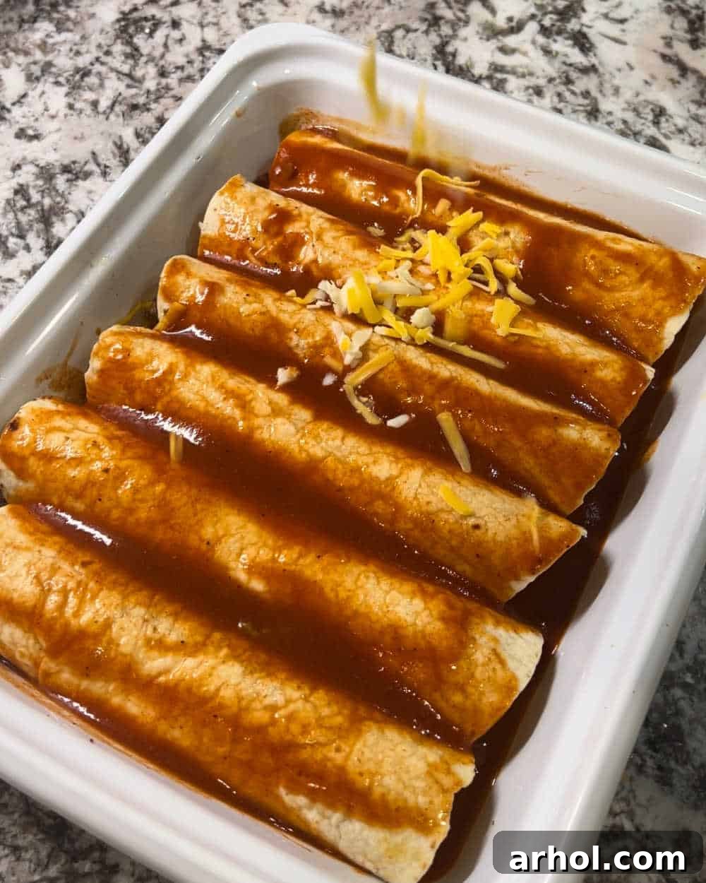 Topping rolled enchiladas with red sauce and additional shredded cheese in a baking dish.