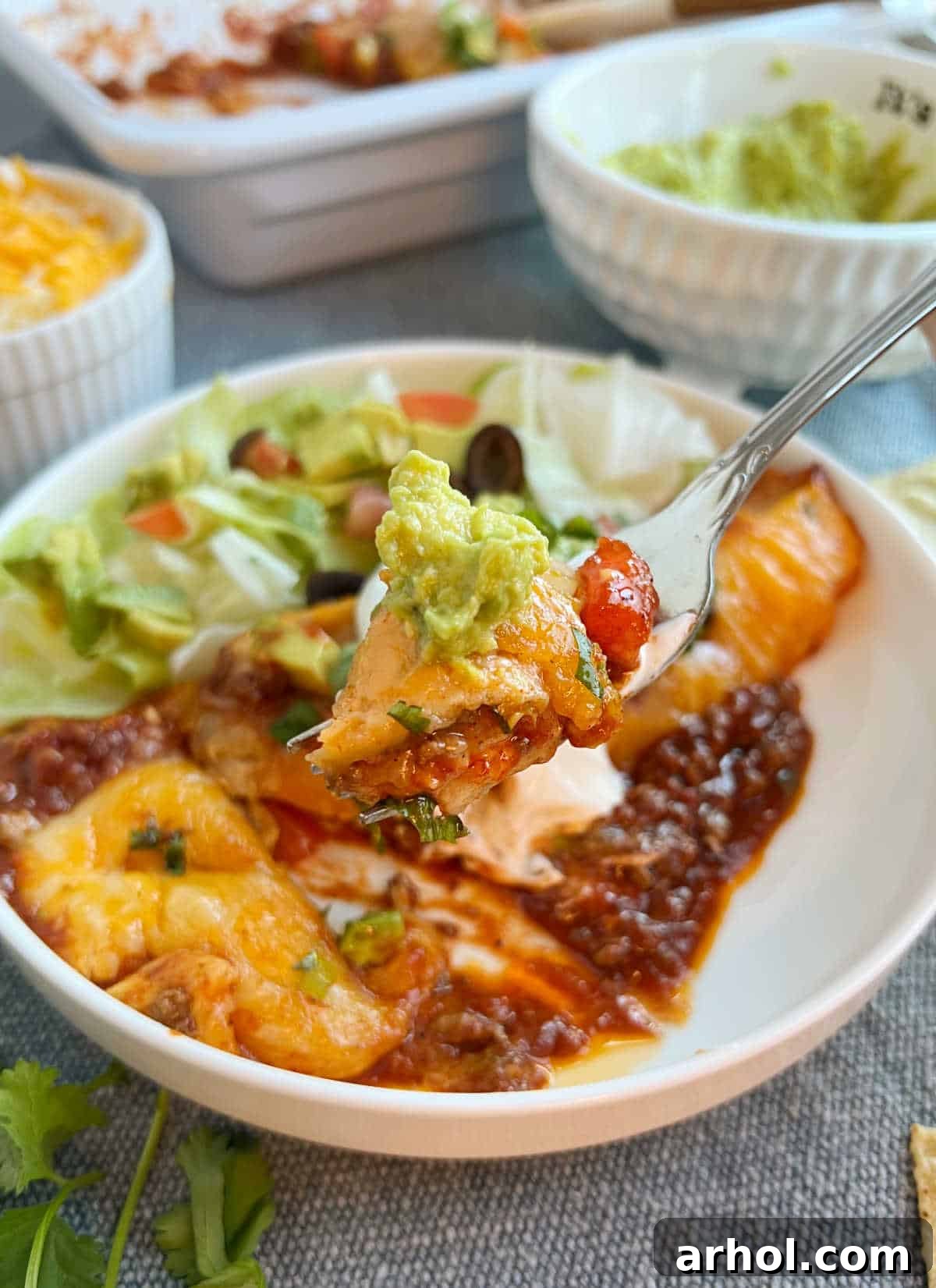 A single ground beef enchilada on a fork with a dollop of guacamole in the background.