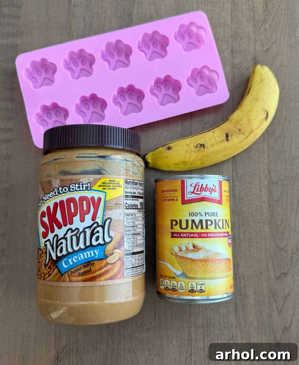 Three core ingredients: peanut butter, ripe banana, and canned pumpkin puree, alongside a paw print treat mold, laid out on a table for making homemade dog treats.