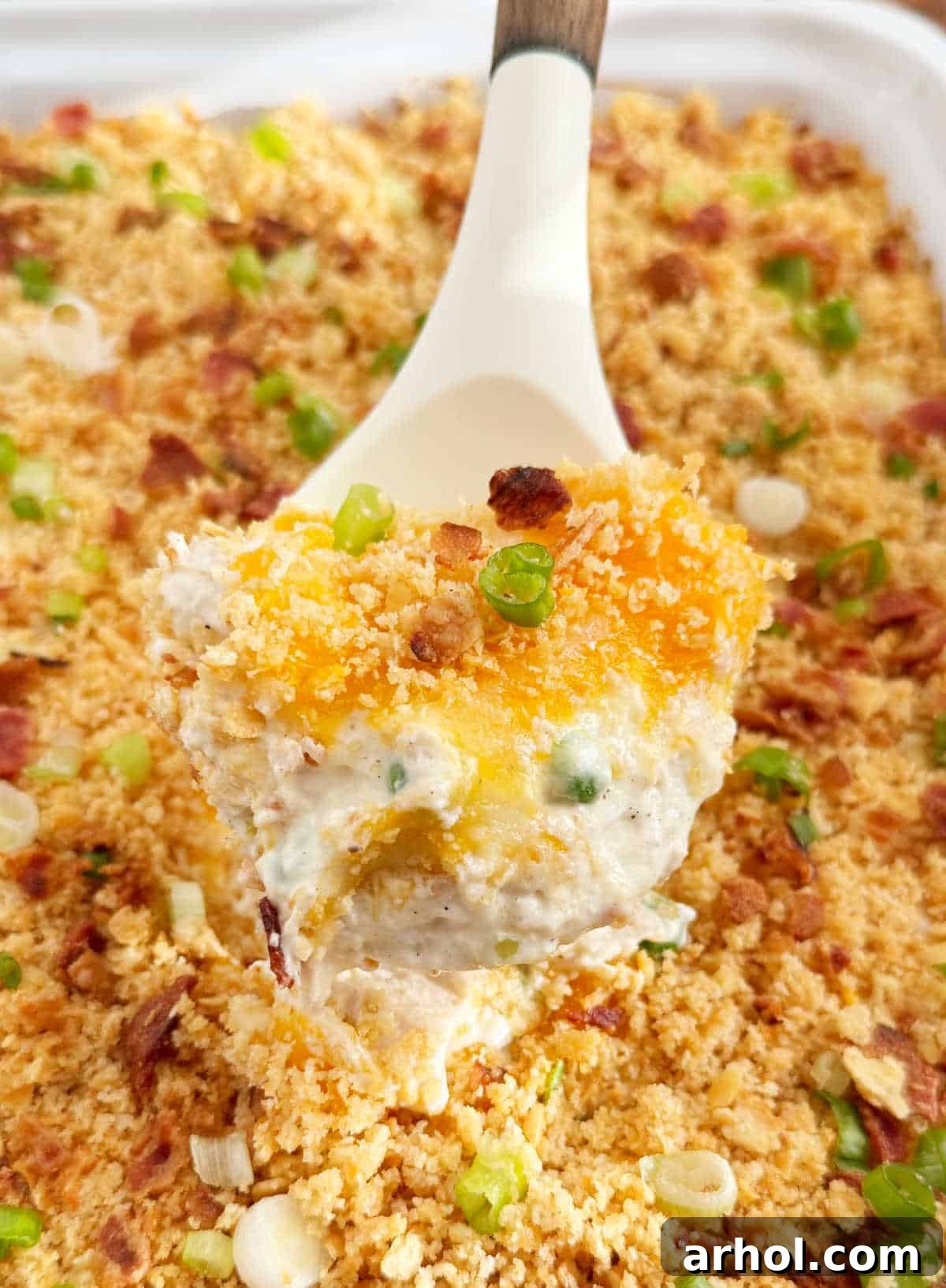 A serving spoon lifting a portion of Million Dollar Chicken Casserole, garnished with fresh green onion, crispy bacon, and bright green peas.