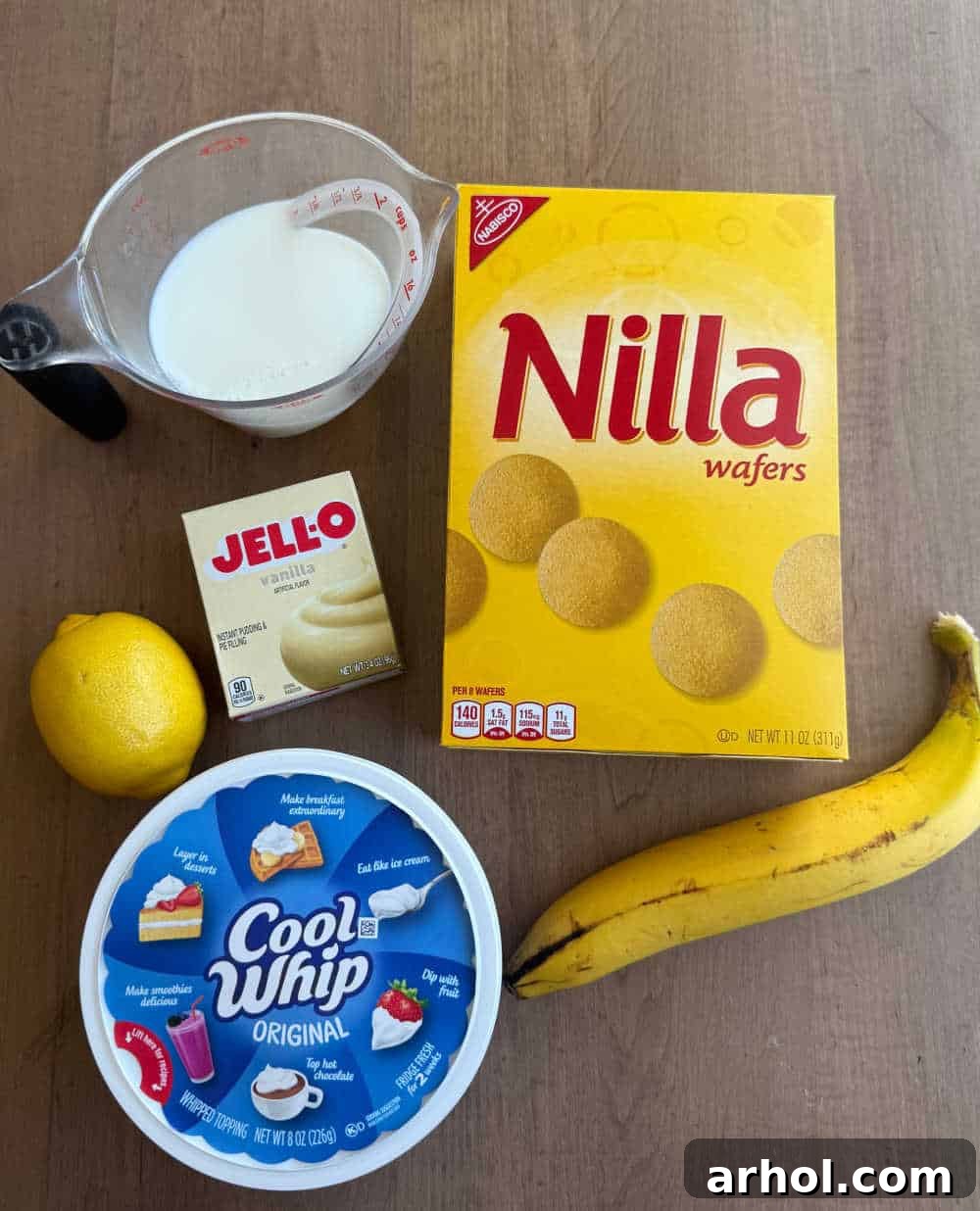 A flat lay photograph showcasing the simple yet essential ingredients for banana pudding dip: a container of Cool Whip, ripe bananas, instant vanilla pudding mix, milk, and a lemon.