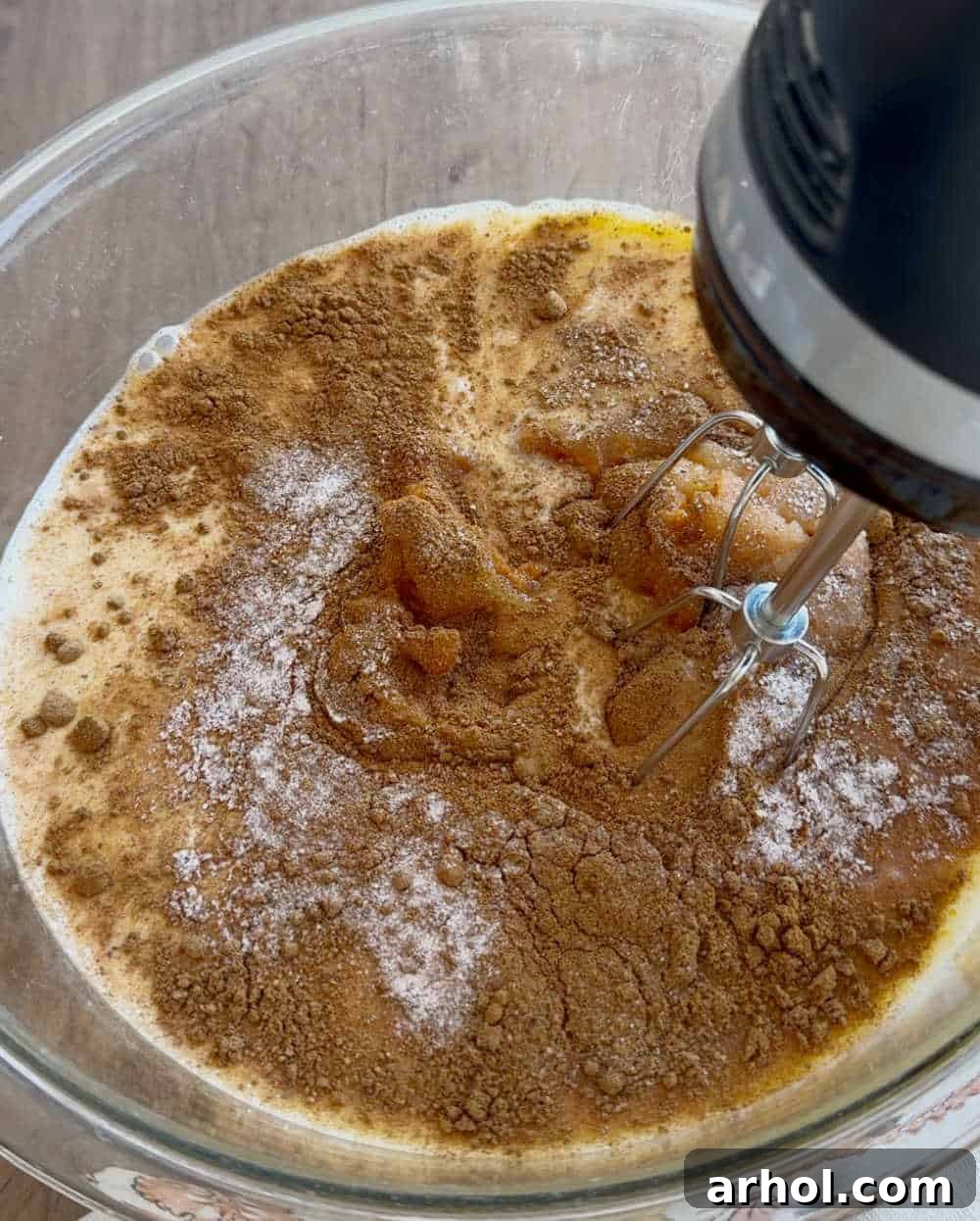 A hand mixer blending pumpkin puree, evaporated milk, eggs, sugar, and spices into a smooth, orange filling in a bowl.