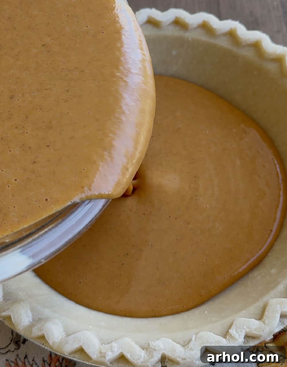 The creamy pumpkin pie filling being poured from a mixing bowl into an uncooked pie crust set in a pie plate.