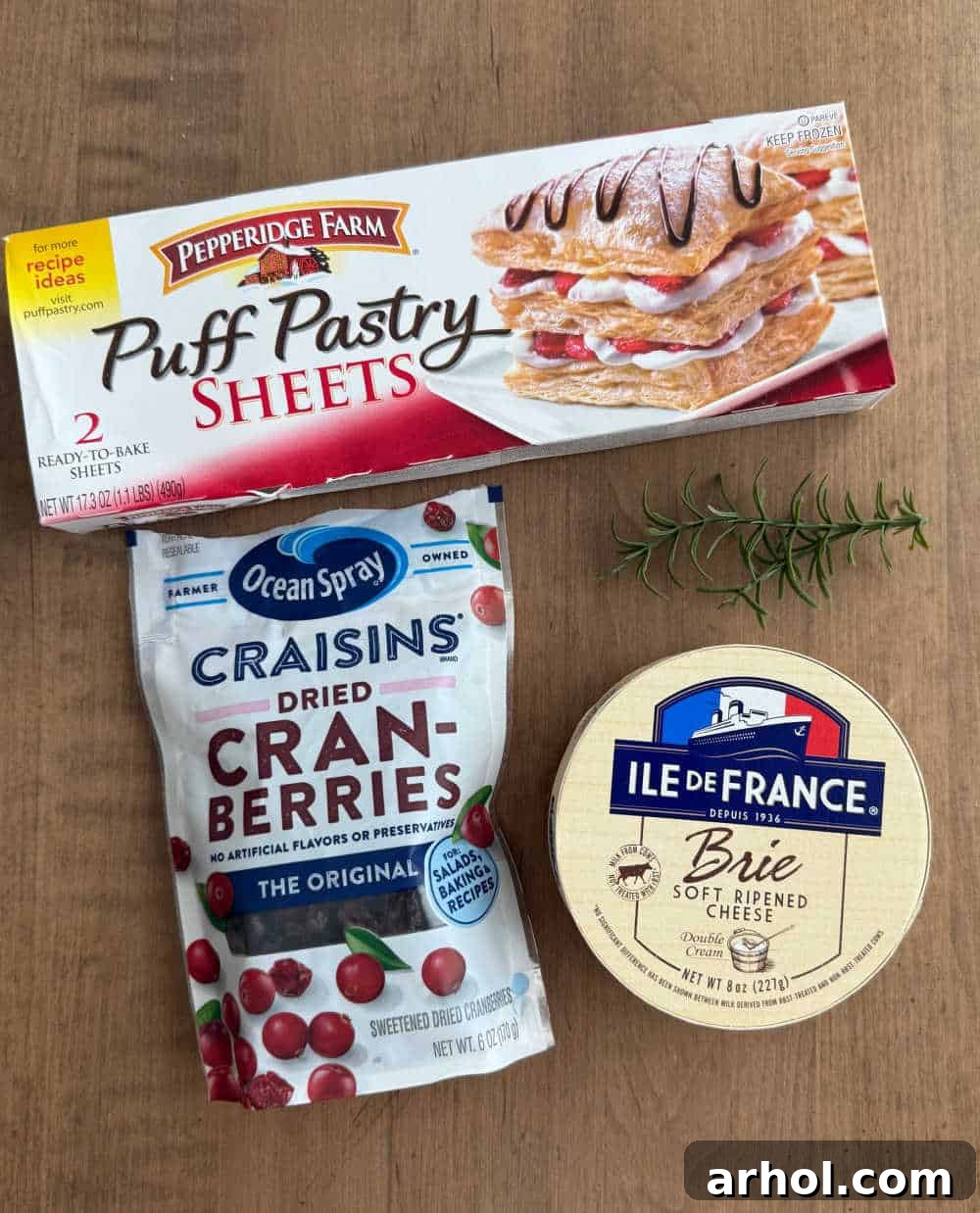 Essential ingredients for cranberry brie bites: a box of puff pastry, Craisins, a wheel of brie, and a fresh rosemary sprig.