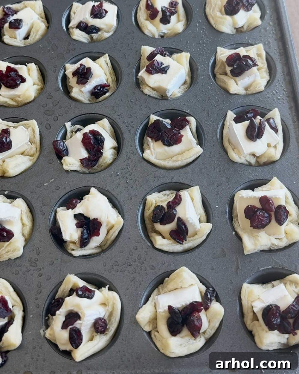 Assembling baked brie bites in a muffin tin with brie cubes and dried cranberries before the second bake.