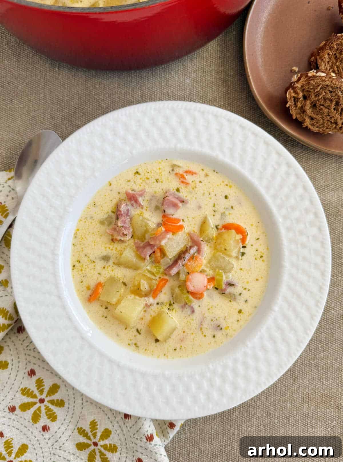 A steaming bowl of creamy ham and potato soup, garnished with fresh parsley and served with a side of crusty bread, featuring visible chunks of ham, tender potatoes, and carrots.