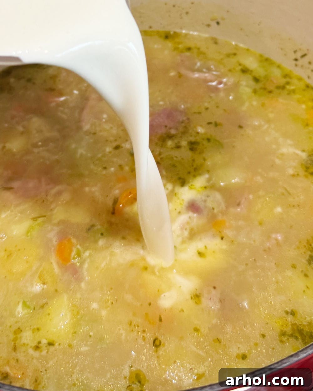 Pouring heavy cream into the simmering ham and potato soup in a Dutch oven for a creamy finish.