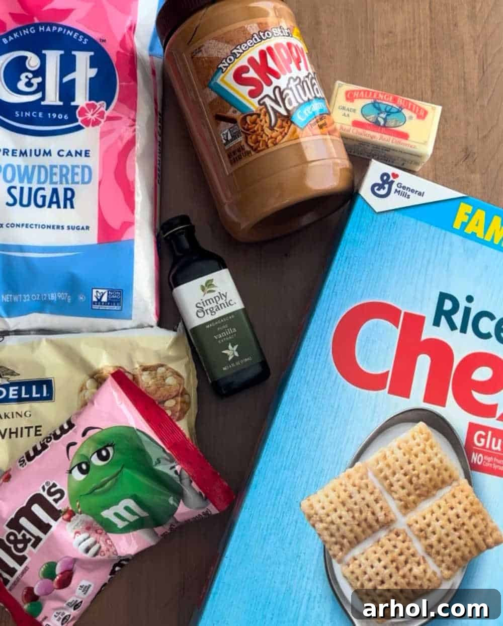 A flat lay showing key ingredients for puppy chow: rice chex cereal, pink and red m&m's, creamy peanut butter, white chocolate chips, butter, and vanilla extract.