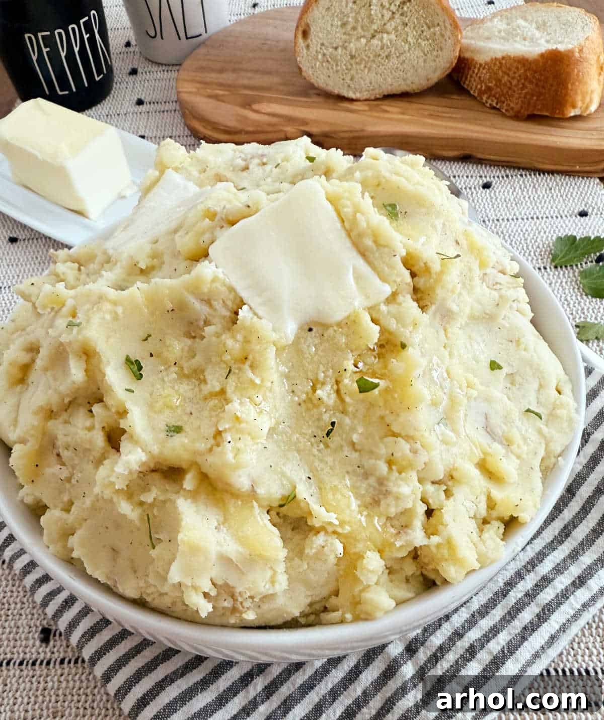 A rustic bowl of mashed potatoes with skin on, topped with a generous pat of melting butter and a sprinkle of fresh herbs. Steam rises gently, highlighting its comforting warmth.