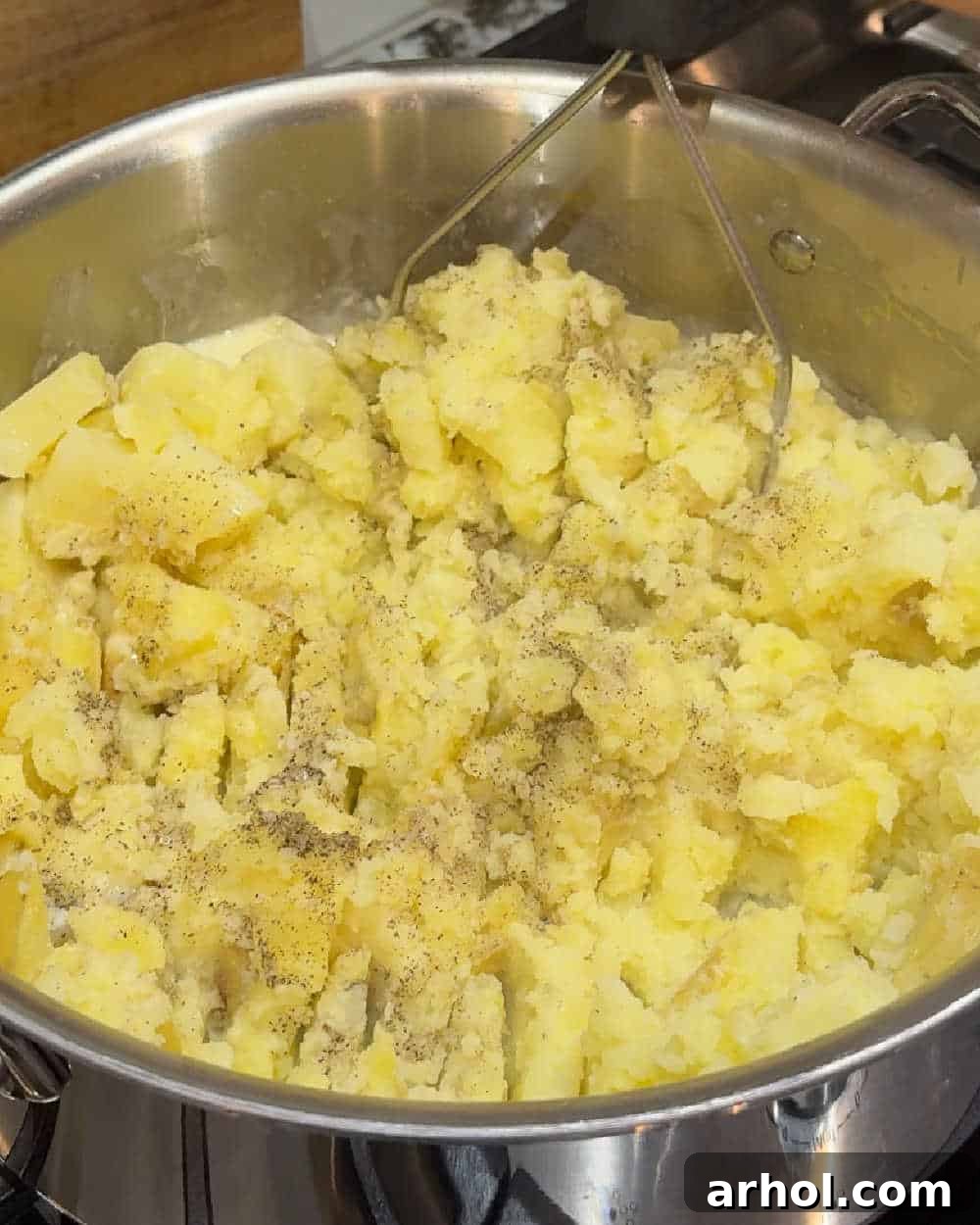 A potato masher actively at work, crushing tender potatoes with skins on inside a large pot, creating a rustic mash.