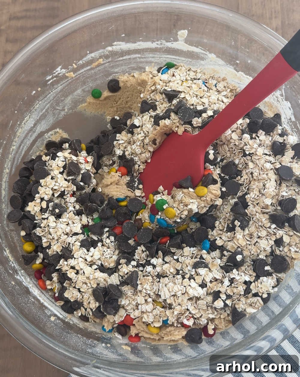 Folding in oats, chocolate chips, and M&M candies into the thick cookie dough mixture in a large bowl.