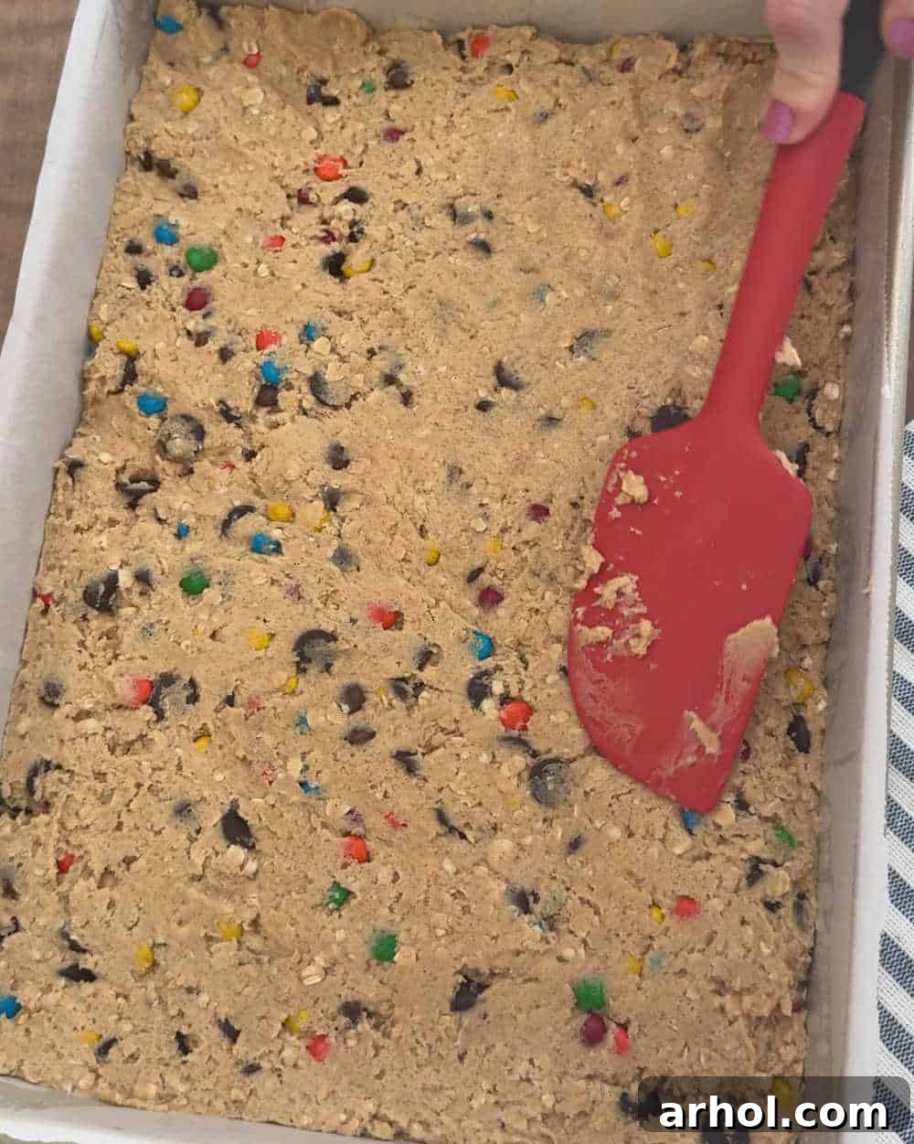 Pressing the thick cookie dough evenly into a prepared baking pan using a rubber spatula.