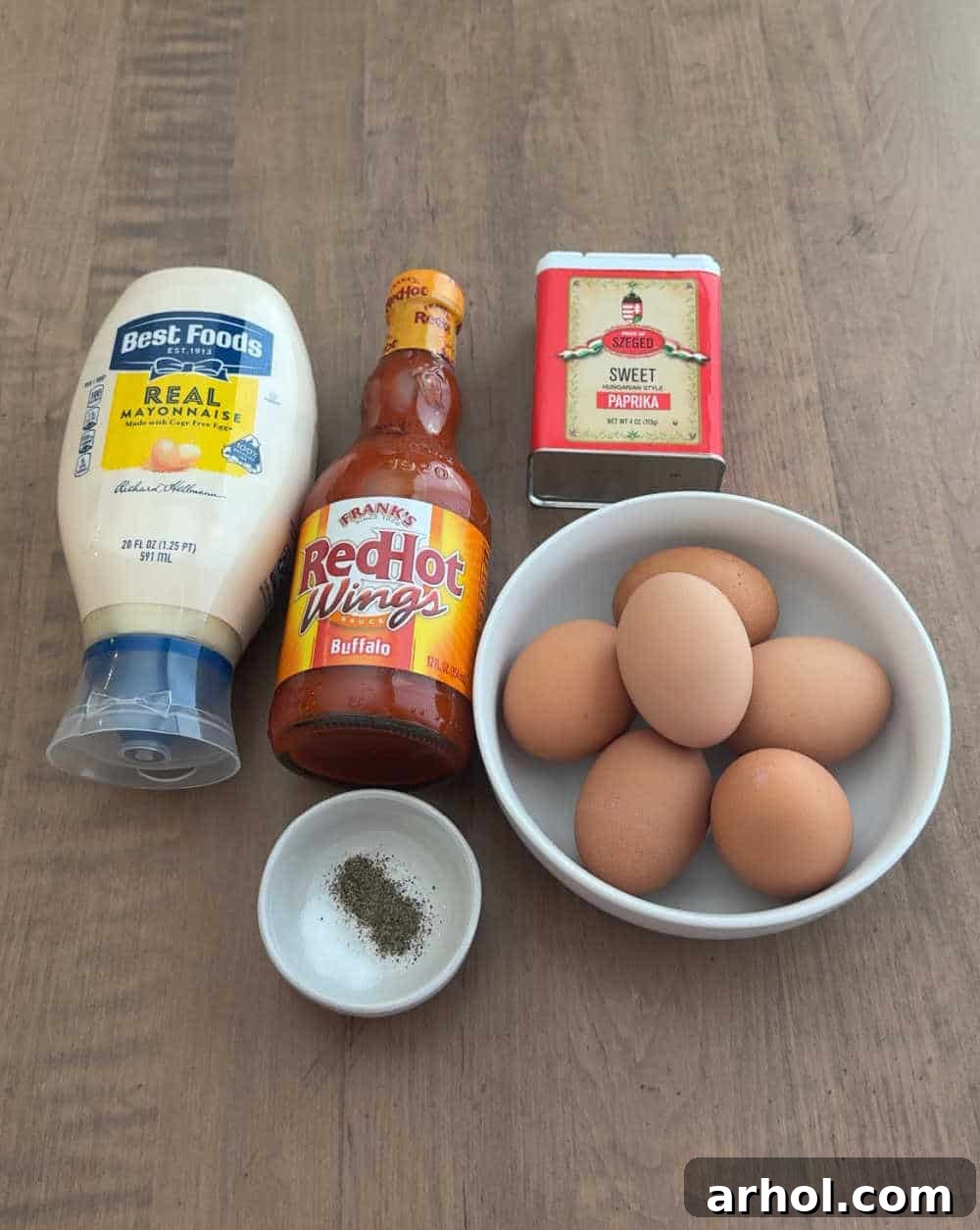 Neatly arranged ingredients for Buffalo deviled eggs: perfectly hard-boiled eggs, a bowl of creamy mayonnaise, a bottle of spicy Buffalo sauce, a small dish of red paprika, and a pinch of salt and pepper.