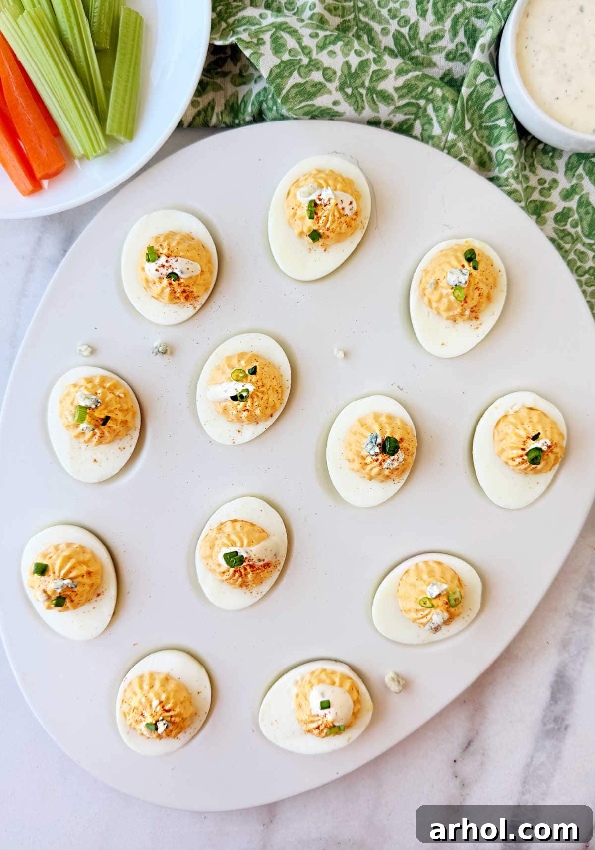 A serving tray filled with Buffalo deviled eggs, some garnished with creamy ranch dressing and others with crumbled blue cheese, artfully arranged alongside fresh carrots and celery sticks.