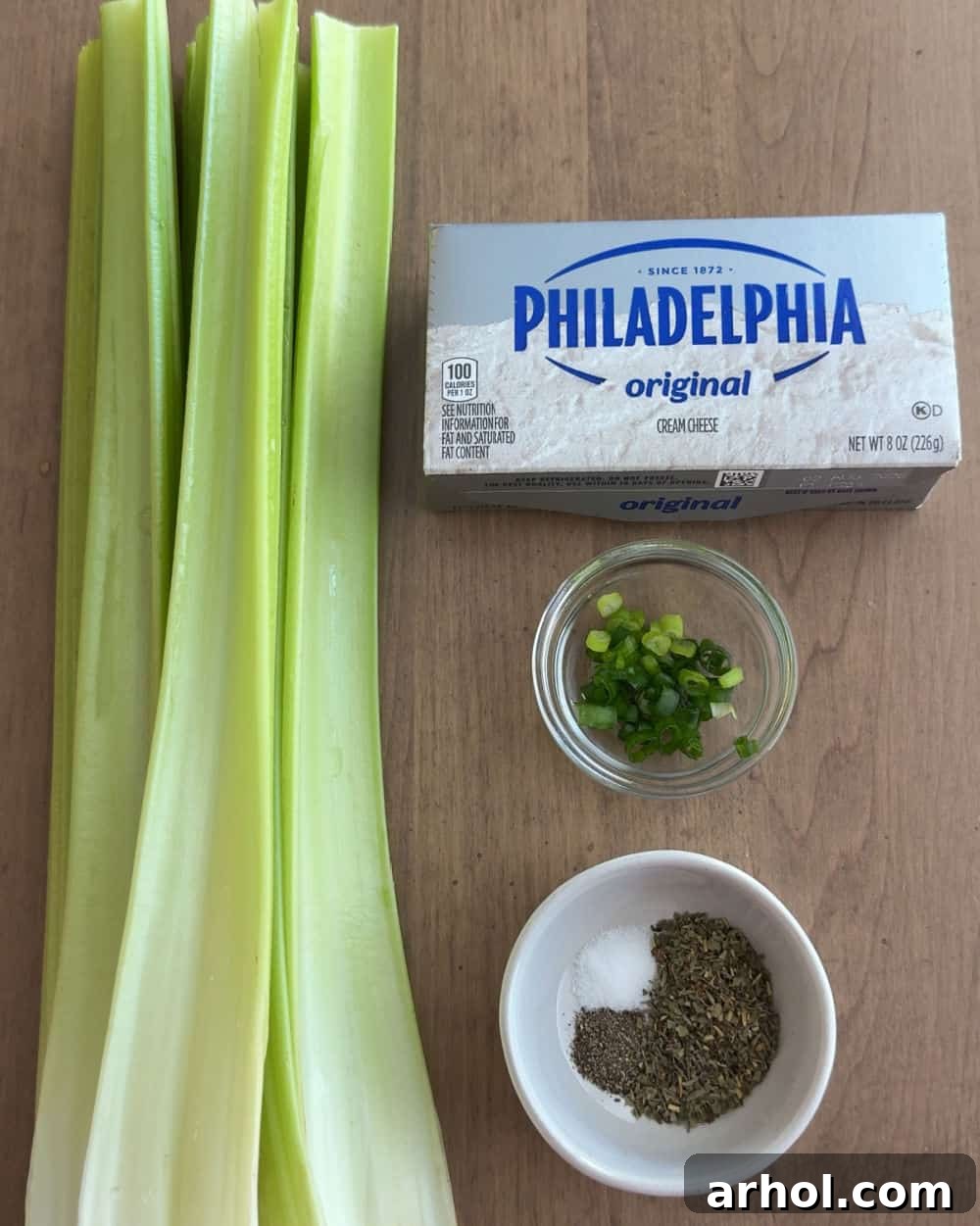 Fresh celery ribs, a block of cream cheese, a small bowl of chopped chives, and various seasonings laid out on a wooden board, ready for preparation.