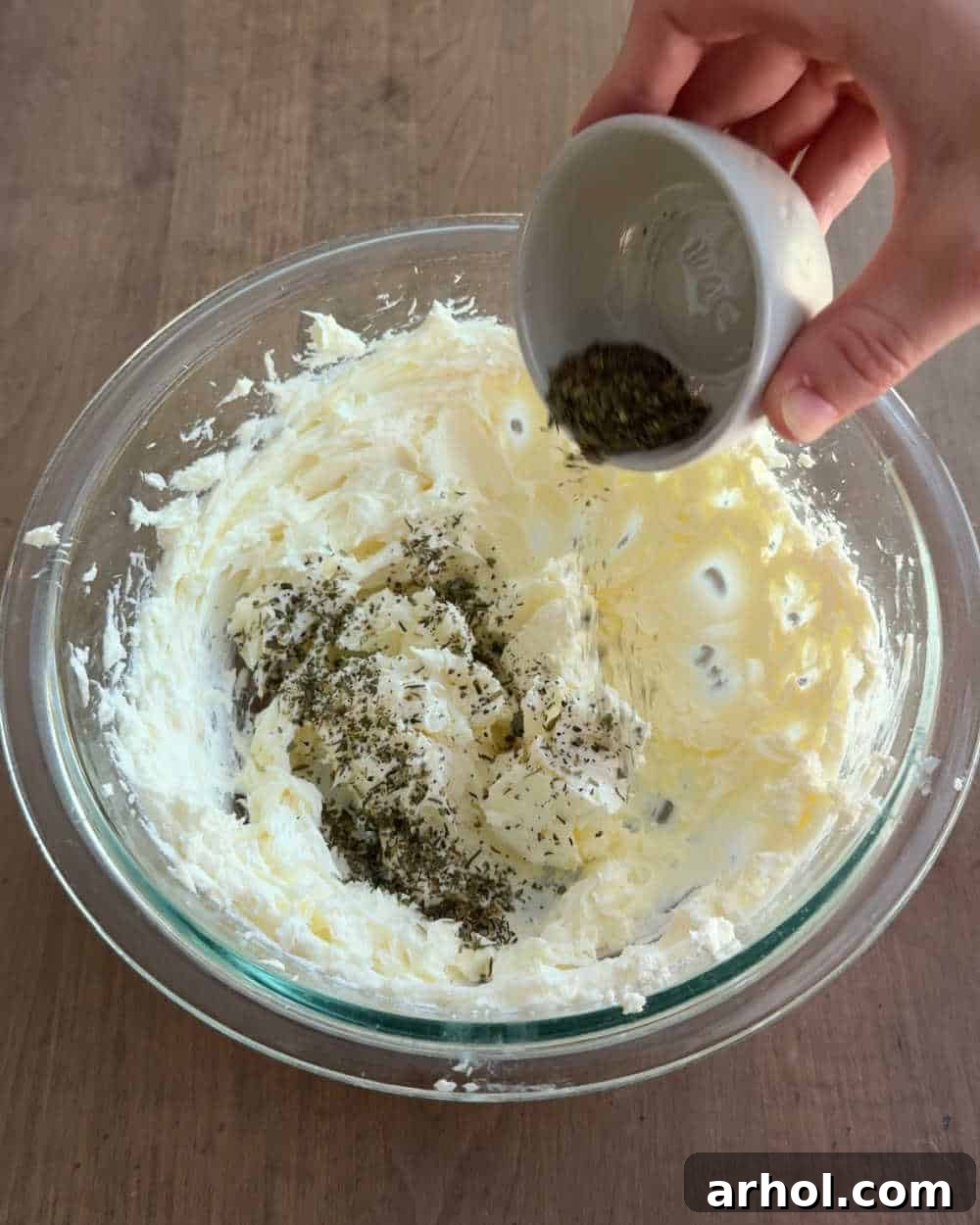 A bowl of softened cream cheese with measuring spoons filled with herbs and spices on top, ready for mixing.