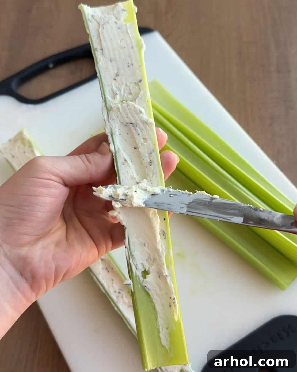 A knife spreading the cream cheese filling smoothly into the curved channel of a celery stalk.
