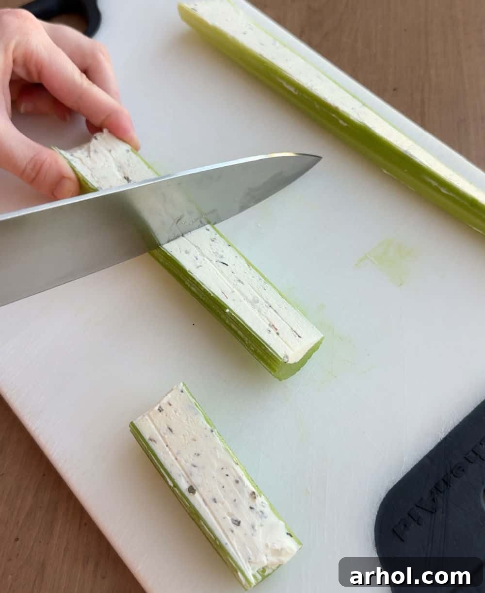 A cutting board with a knife slicing a cream cheese-stuffed celery stalk into bite-sized pieces.