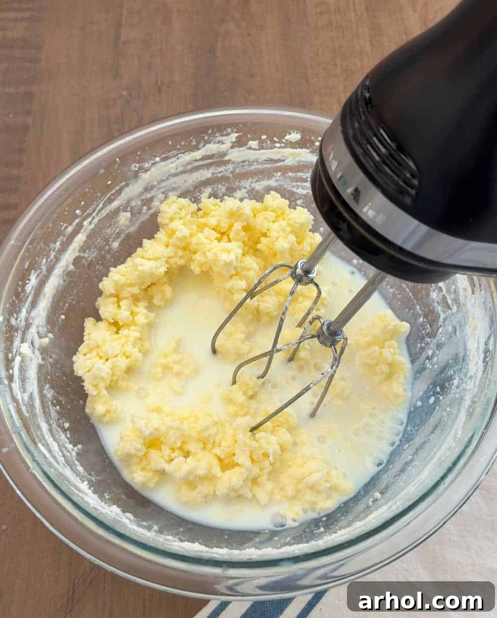 An electric mixer actively turning heavy cream into butter in a large mixing bowl, showing the transition from liquid to solid.