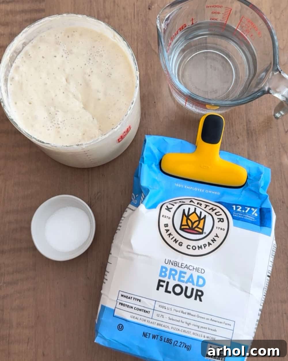 Ingredients for same day sourdough bread: a bowl of bread flour, active sourdough starter in a jar, a bowl of sea salt, and a measuring cup of warm water.