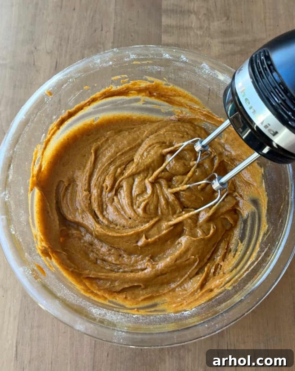 A close-up shot of pumpkin batter being mixed in a large bowl with an electric hand mixer, showing the rich orange color and smooth consistency.