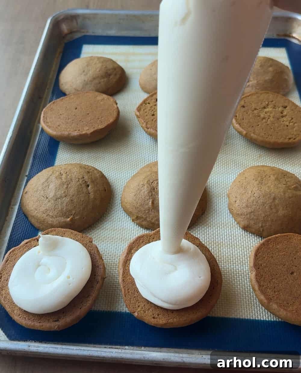 Hands carefully piping a generous amount of cream cheese filling onto the flat side of a cooled pumpkin cookie, using a piping bag.