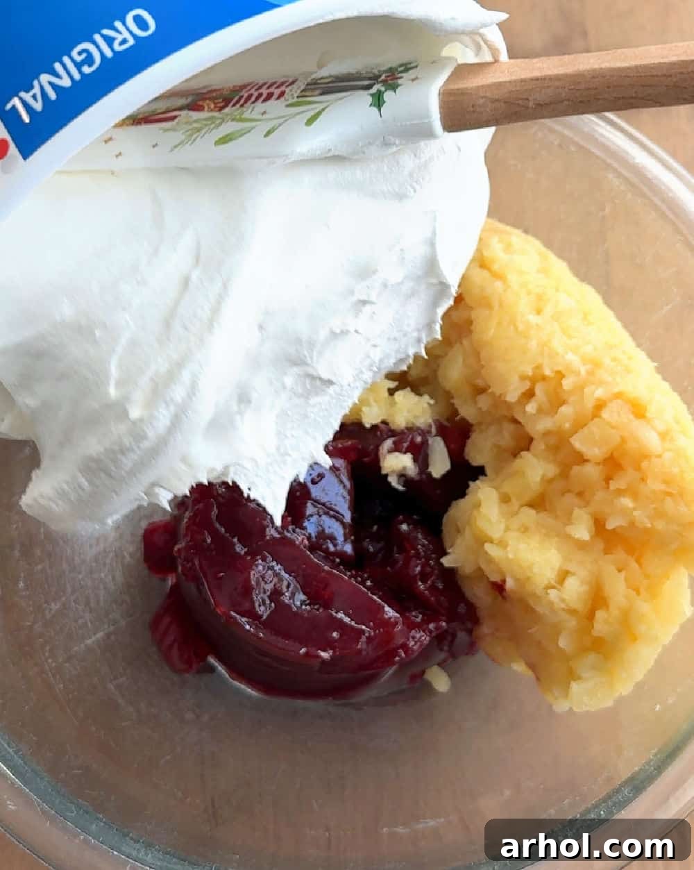 Adding a generous dollop of creamy, thawed whipped topping (Cool Whip) into the vibrant cranberry and pineapple mixture using a spatula.