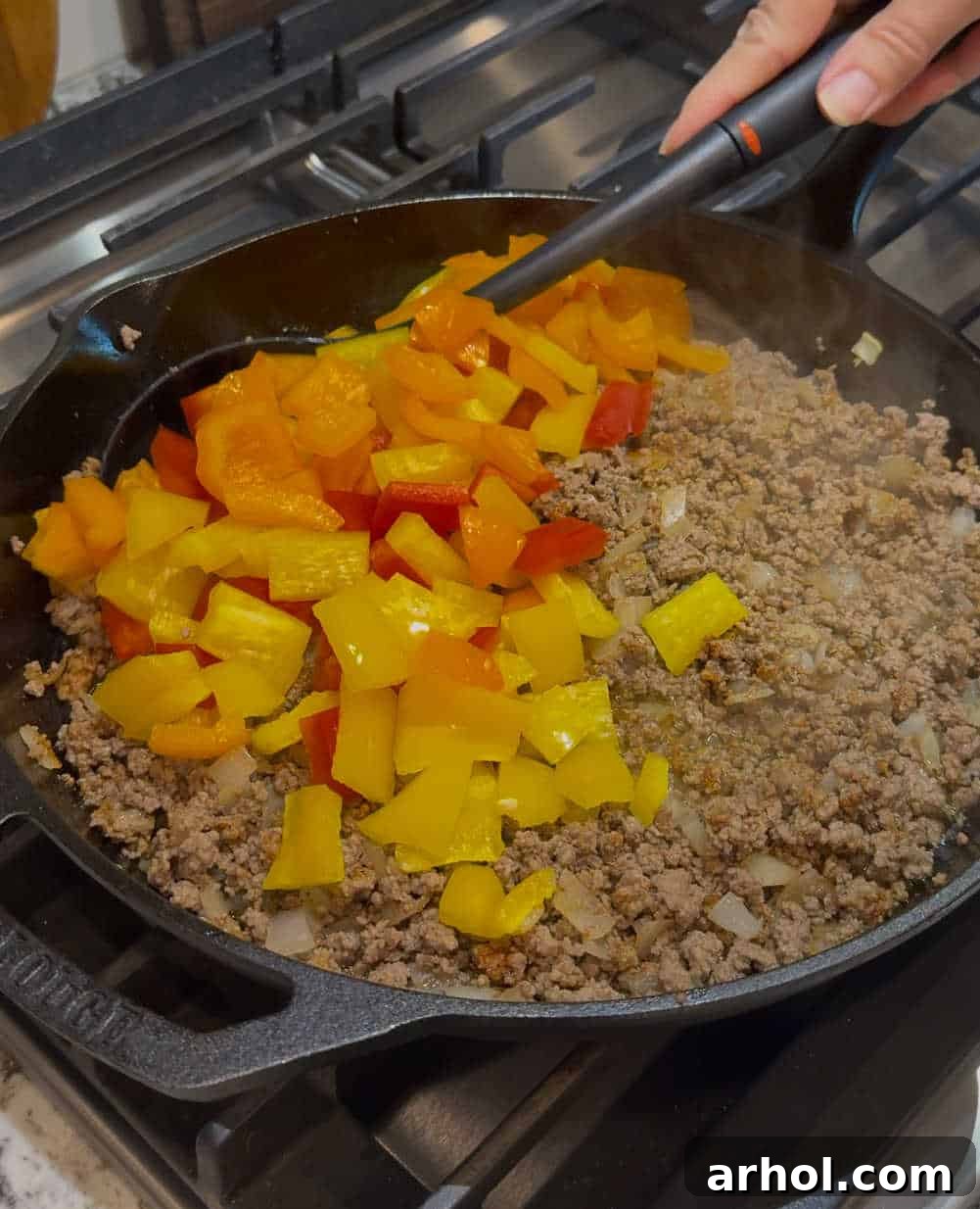 Chopped bell peppers being stirred into ground beef and onions in a large skillet, beginning to soften.