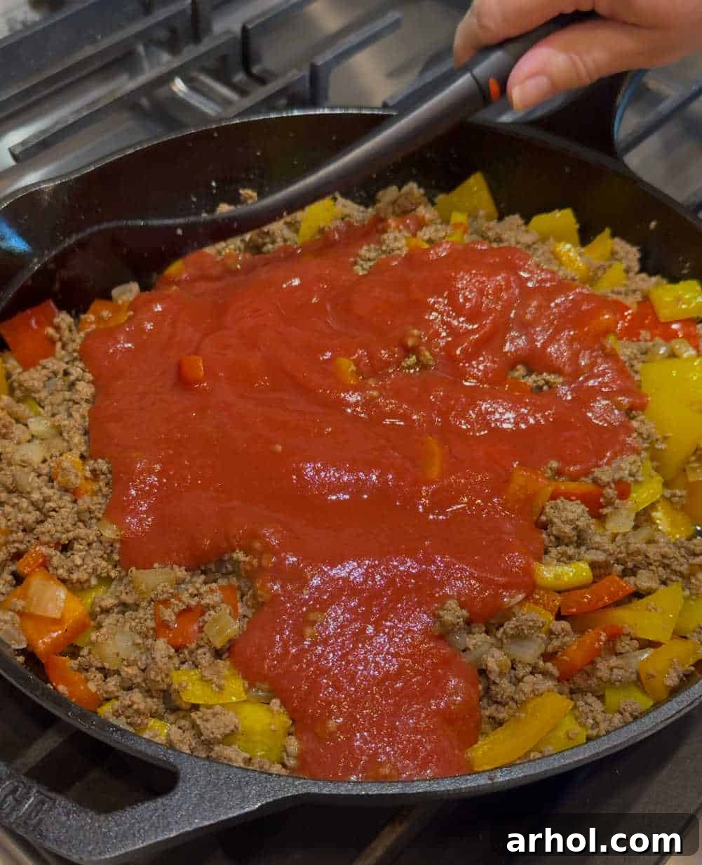A can of tomato sauce being stirred into the cooked ground beef and bell pepper mixture in a skillet, creating a rich, red base.