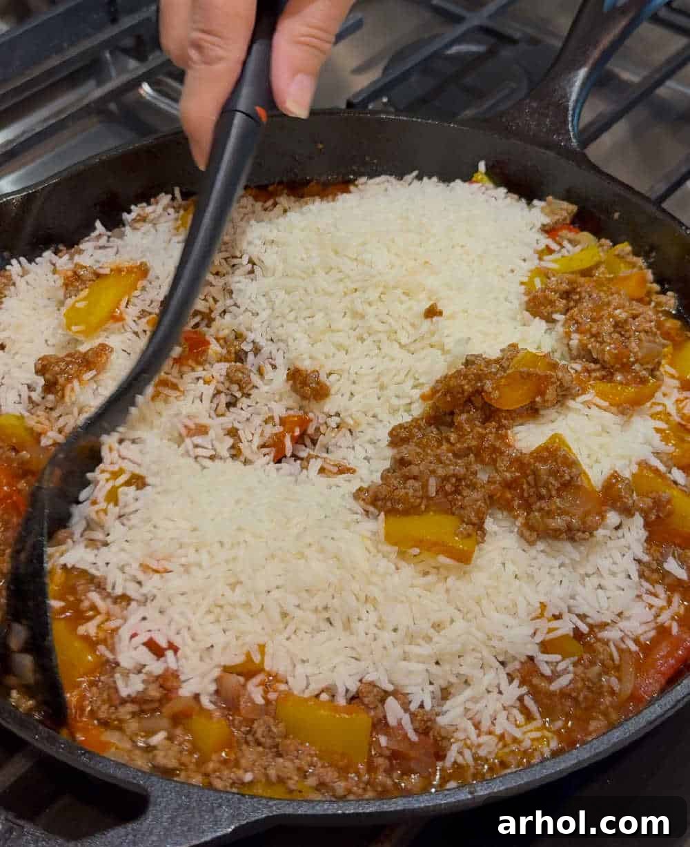 Instant rice being stirred into the tomato sauce, ground beef, and pepper mixture in a large skillet, before adding broth.