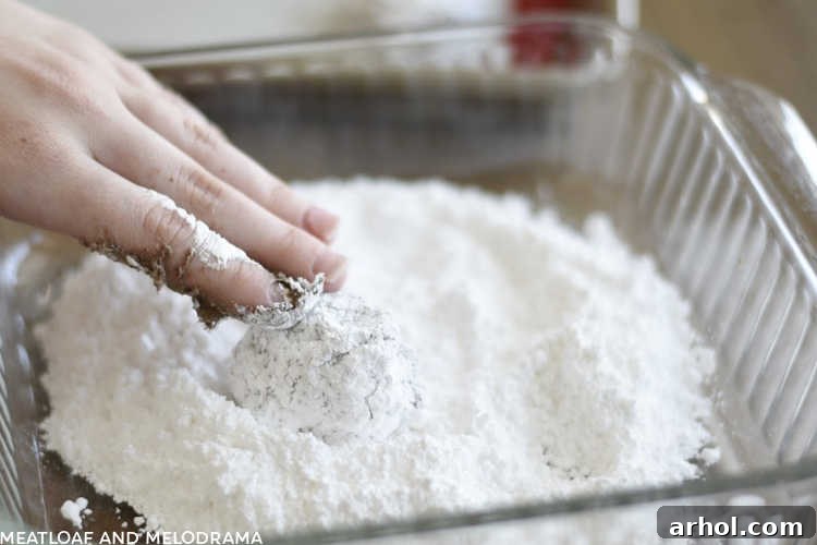 A hand rolling a chocolate cookie dough ball generously in a bowl of powdered sugar.