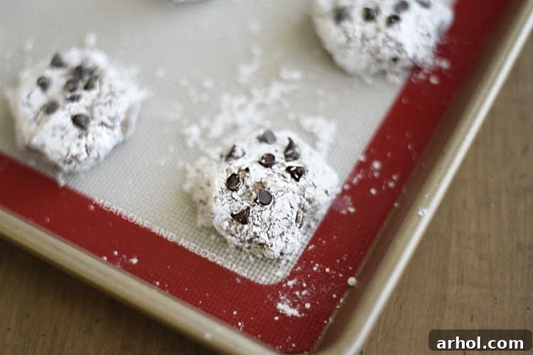 Chocolate crinkle cookies, covered in powdered sugar and topped with mini chocolate chips, arranged on a baking sheet ready for the oven.