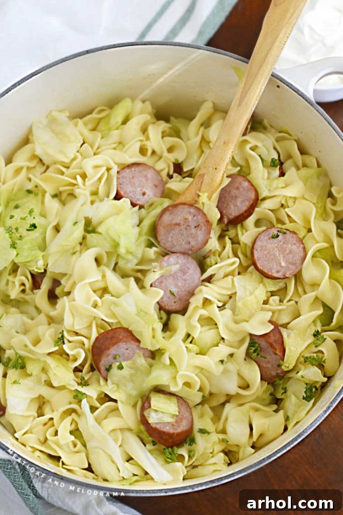 A wooden spoon rests in a white Dutch oven filled with Haluski and Kielbasa, showcasing the fried cabbage, egg noodles, and smoked sausage, ready to be served.