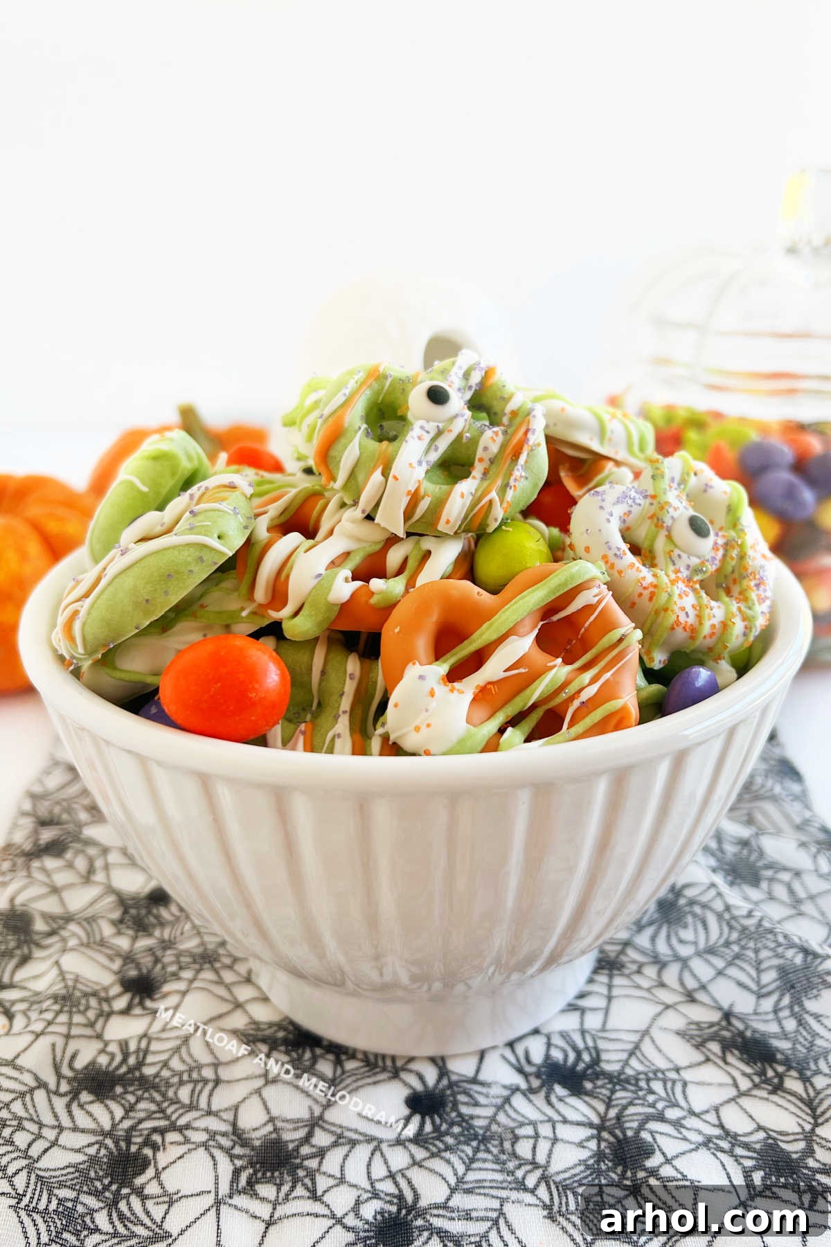 A festive bowl of Halloween pretzels, brightly dipped in orange, green, and white candy coating, adorned with playful candy eyes and surrounded by an assortment of Halloween candies.