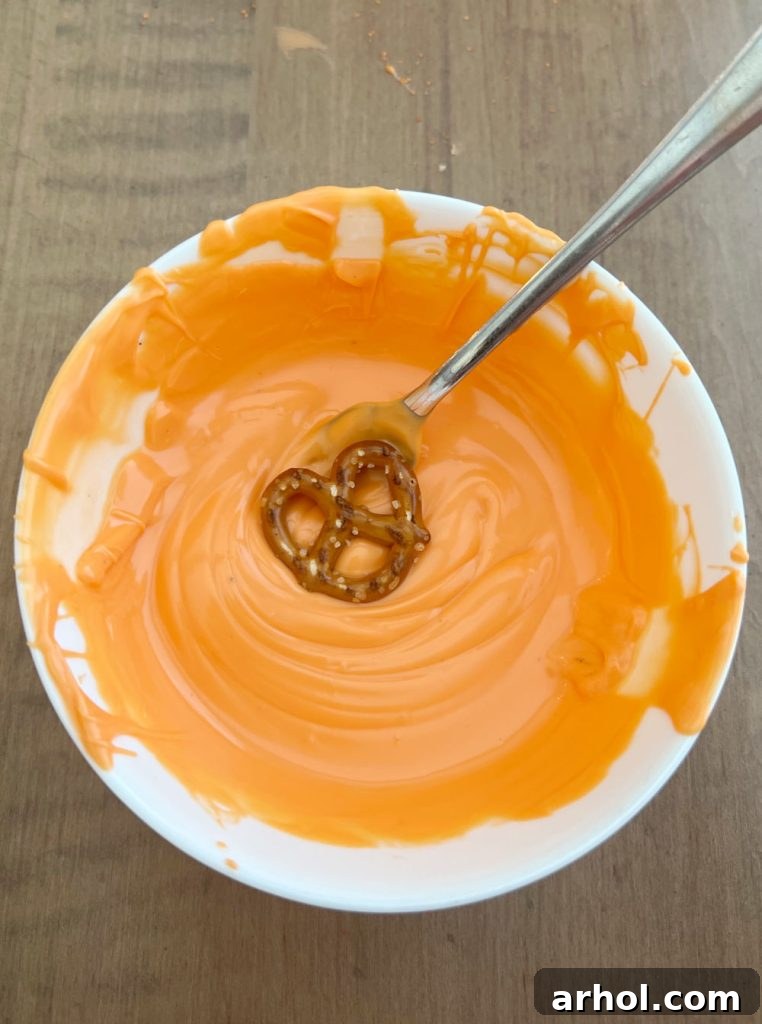 A mini pretzel twist being carefully dipped into a bowl of bright orange melted candy coating using a fork, with excess dripping off.
