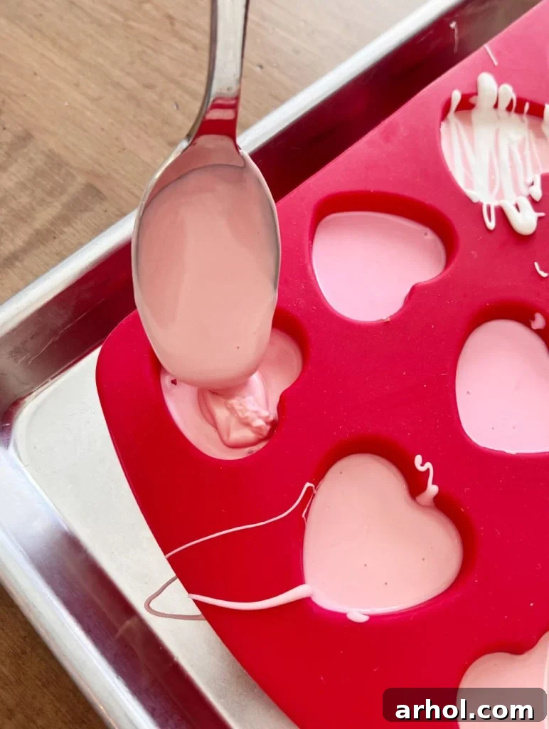 Melted pink candy coating being poured into a heart-shaped silicone mold, with sprinkles visible in some compartments.