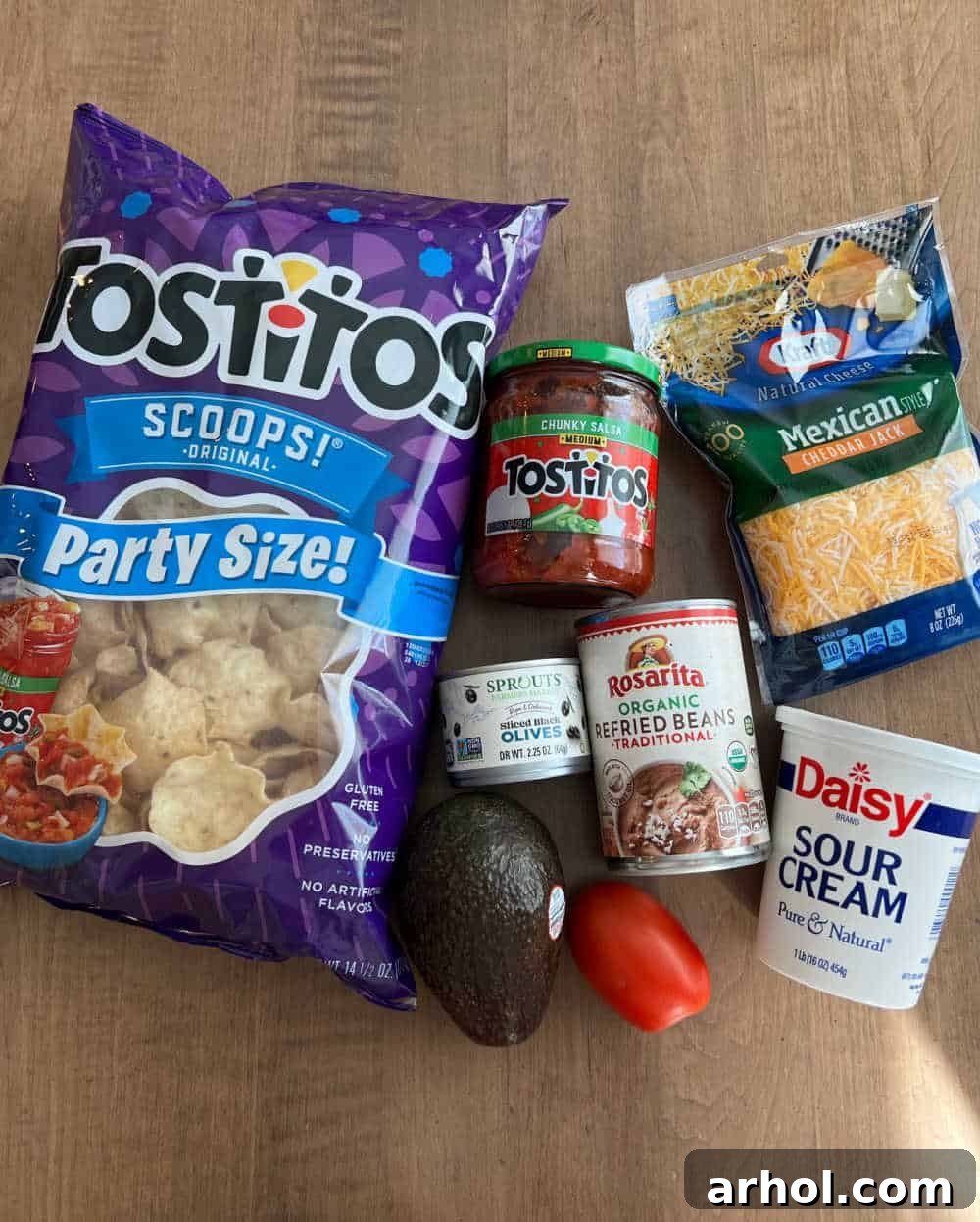 All the fresh ingredients for a seven-layer dip laid out: refried beans, avocado, sour cream, salsa, cheese, tomato, olives, and a bag of tortilla scoops.