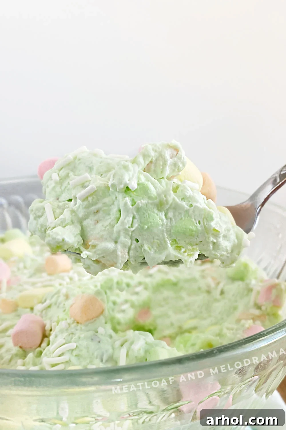 A serving spoon lifting a spoonful of vibrant green watergate salad, revealing colorful fruit-flavored marshmallows and pineapple chunks, ready to be enjoyed.