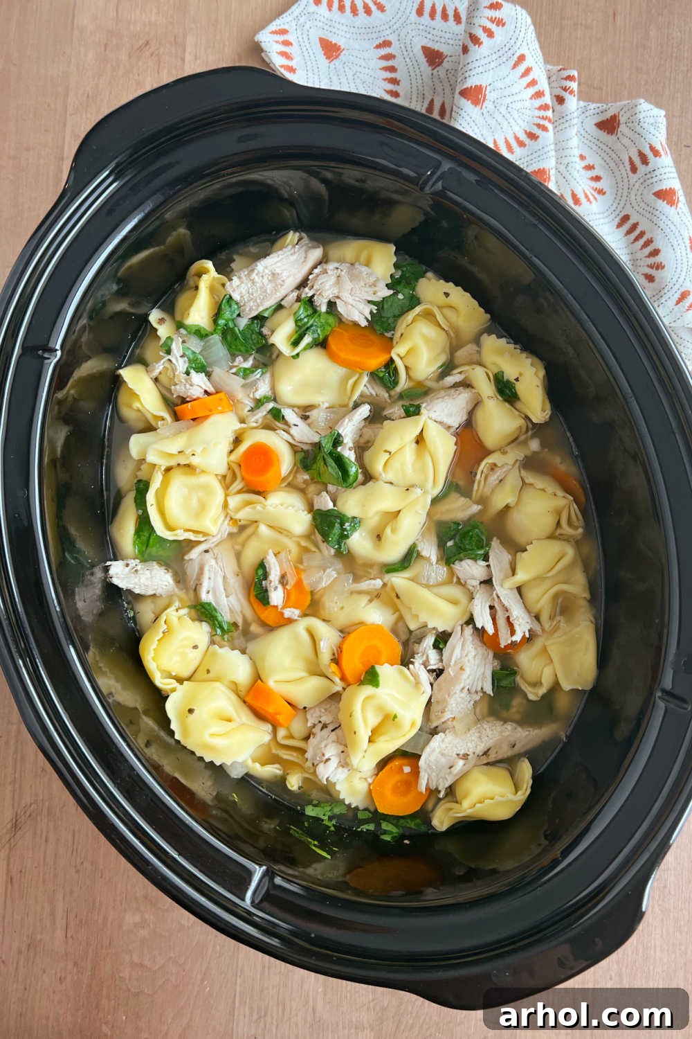 A steaming bowl of Crock Pot Chicken Tortellini Soup with shredded chicken, carrots, and fresh spinach in a slow cooker ladle.