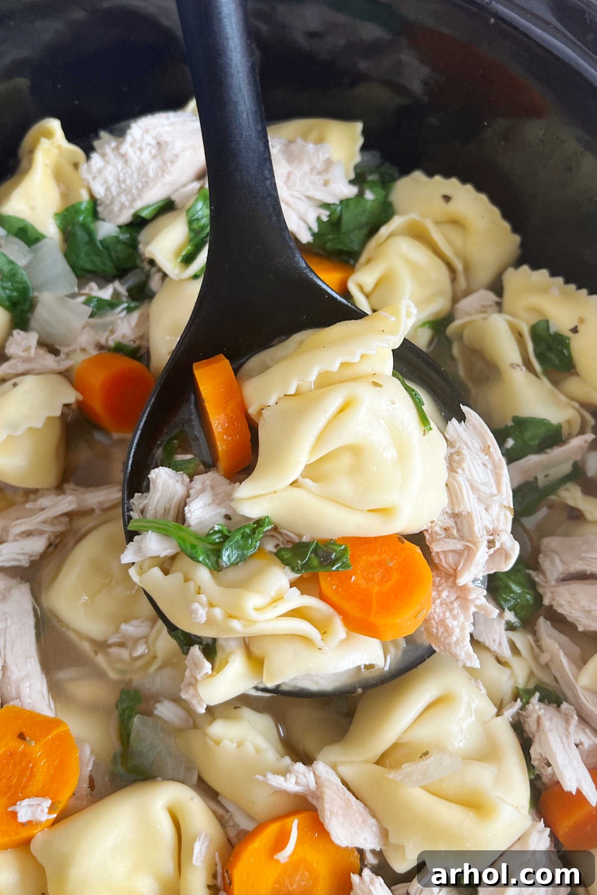 A ladle full of slow cooker chicken tortellini soup being served from the crock pot.