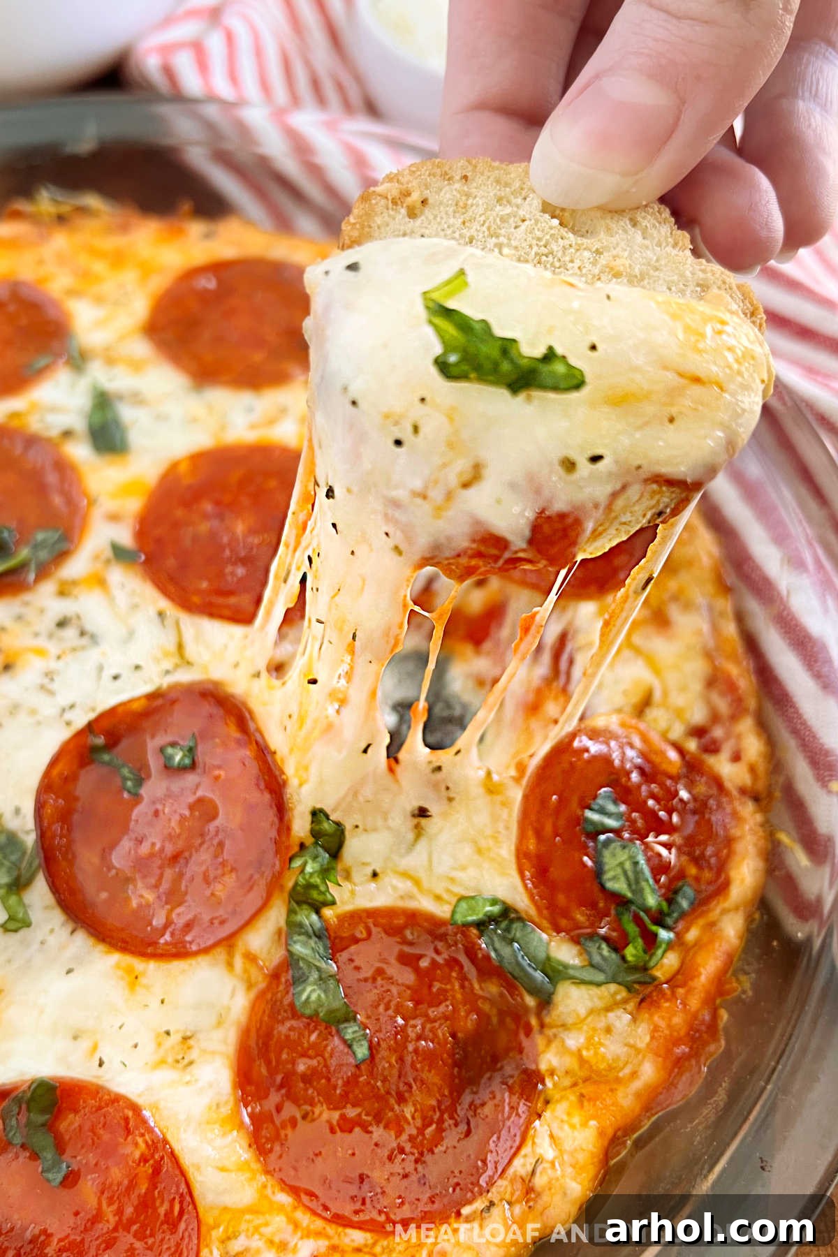 Hand dipping toasted bread round into cheesy pepperoni pizza dip, showcasing melted cheese and crispy pepperoni in a pie dish.
