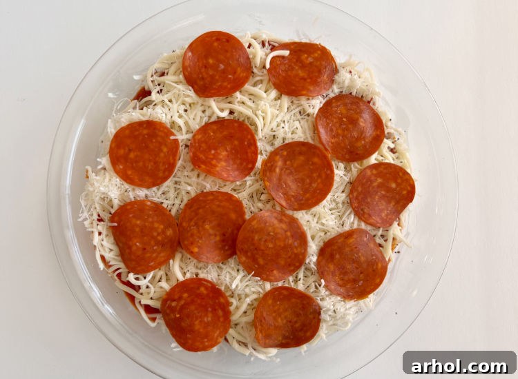 Unbaked pepperoni pizza dip in a pie plate, with all layers assembled, generously topped with pepperoni slices.